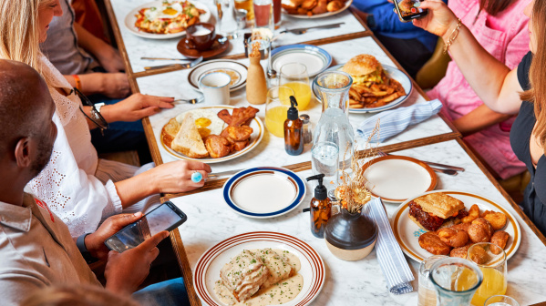 Overhead photo of a group of people gathered around a restaurant table filled with breakfast and brunch dishes. Plates include eggs and toast, bacon, a burger with fries, a sauced entrée, and other shared items, along with drinks like orange juice and water. Several people are holding phones and taking photos of the food.