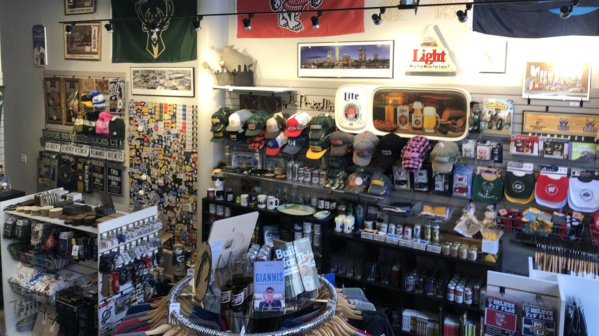 Interior of a Milwaukee-themed retail shop filled with local sports and city merchandise. Shelves and wall displays are packed with baseball caps, glassware, mugs, magnets, and souvenirs featuring teams like the Bucks and Brewers. Framed posters, vintage signs, and banners line the upper walls, while a central display table holds books and additional items. The space is brightly lit and densely arranged, creating a colorful, curated storefront focused on Milwaukee pride.