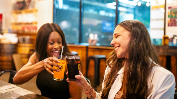 Two women smiling and clinking glasses of Sprecher soda at a wooden table inside a brewery