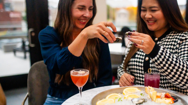Two women smiling and toasting with oysters at a table featuring a seafood platter and cocktails, seated indoors with soft natural lighting.