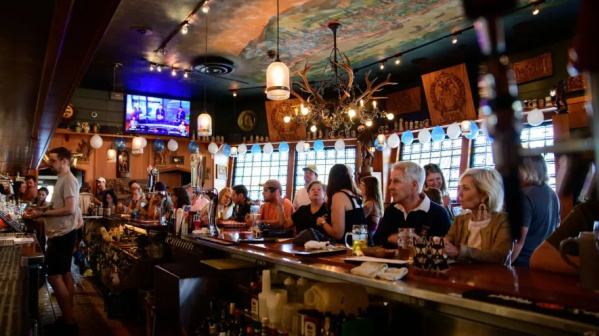 A lively bar scene with guests seated shoulder to shoulder, enjoying drinks beneath warm lighting, festive décor and a painted ceiling that adds character and energy to the space.