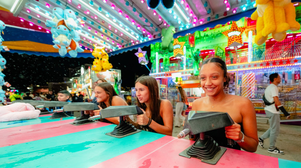 Three women smile while competing at a carnival squirt gun game beneath colorful lights and oversized stuffed animal prizes at the Wisconsin State Fair.