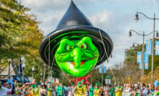 A large witch blowup parade float is pictured with a green face and black witch's hat. Several people are holding ropes that anchor the float.