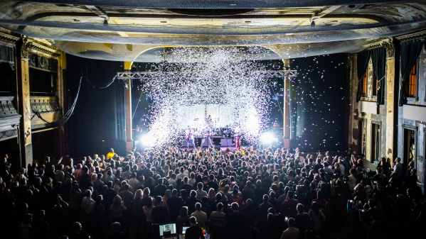 Interior view of Turner Hall ballroom during a live concert, showing a packed crowd of hundreds of people facing an illuminated stage. The historic venue's ornate architecture is visible with decorative columns, balconies, and detailed ceiling work. Dramatic stage lighting creates bright white beams cutting through the dark space, with confetti or snow effects falling from above.