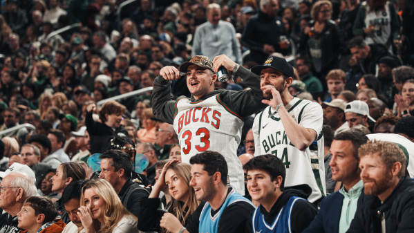 Two excited fans wearing Milwaukee Bucks jerseys cheer from their seats during a packed game at Fiserv Forum, surrounded by a crowd of fellow spectators watching the action.