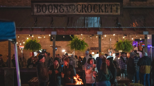 A crowd gathers at Boone & Crockett for an outdoor event at night. People in flannel and jackets socialize with drinks around a fire pit under string lights and hanging plants. The rustic bar sign hangs above the scene, creating a cozy festival atmosphere.