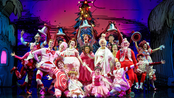 A large group of actors in colorful, whimsical costumes pose energetically on stage in front of a festive, cartoon-style backdrop, likely from a holiday-themed theater production inspired by Dr. Seuss.