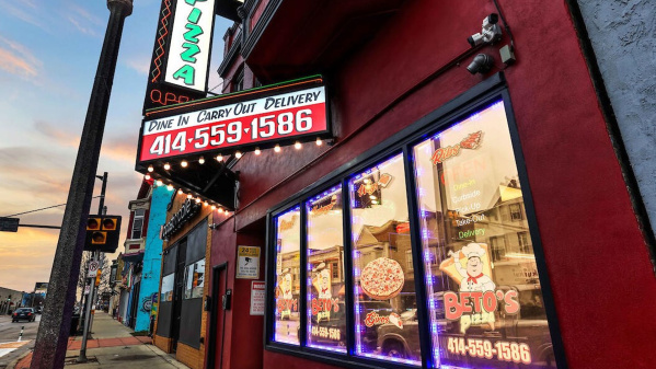 Street-view of Beto’s Pizza, a brightly lit red storefront in Milwaukee with neon signs advertising dine-in, carry-out, and delivery, including the phone number 414-559-1586. The building glows at dusk with colorful signage and reflections in the windows.