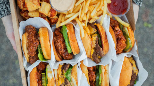 A person holding a box filled with cheeseburgers, fried chicken sandwiches, French fries, tater tots, and dipping sauces.