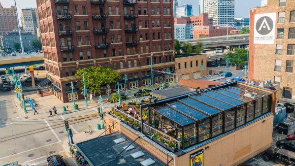 The Lux Skyhaus at Cafe Benelux, a glass greenhouse rooftop dining space in Milwaukee, overlooks a pedestrian plaza with teal lamp posts. The transparent structure reveals lush greenery and diners inside, surrounded by historic red brick buildings and downtown Milwaukee's skyline.