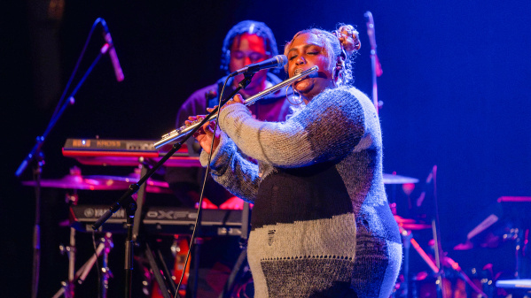 Musician playing the flute and singing into a microphone on stage, with a keyboardist and drum set in the background under blue and purple stage lights.
