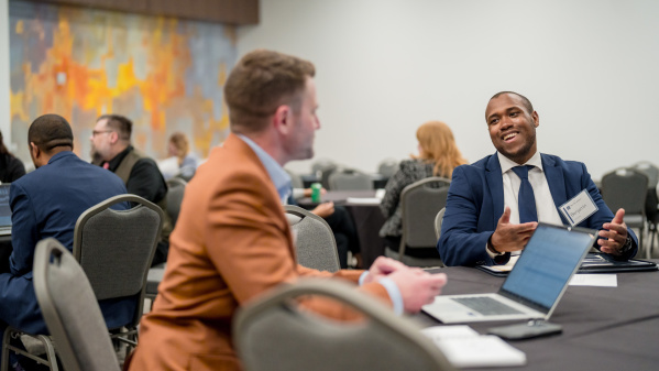 Two business professionals conversing at a conference table - one in an orange blazer with back to camera, the other in a navy suit smiling and gesturing while speaking. Multiple attendees visible in background conference room setting.