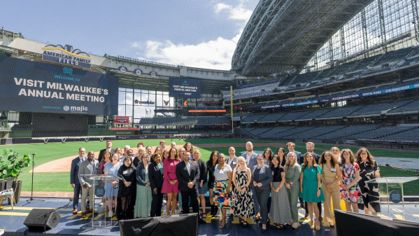 Group photo of attendees at Visit Milwaukee's Annual Meeting, posed on a stage at American Family Field (Milwaukee Brewers stadium) with stadium seating and banners visible in the background.