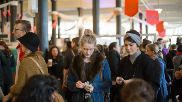 A busy indoor market scene with people browsing and making purchases. Two women in the foreground count money, while crowds of shoppers fill the background among vendor booths and hanging signs.