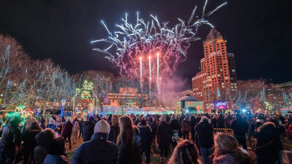 A crowd gathers in downtown Milwaukee at night to watch a dazzling fireworks display lighting up the sky above city buildings, surrounded by festive holiday lights and winter decorations.