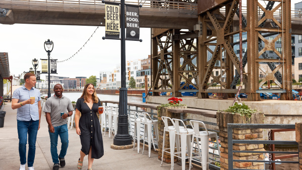 Three people walk along the Milwaukee Riverwalk outside Lakefront Brewery, holding beers and smiling. They pass under an industrial bridge structure with banners reading “Cold Beer in Here” and “Beer. Food. Beer.” while white bar stools and flower planters line the railing overlooking the river.