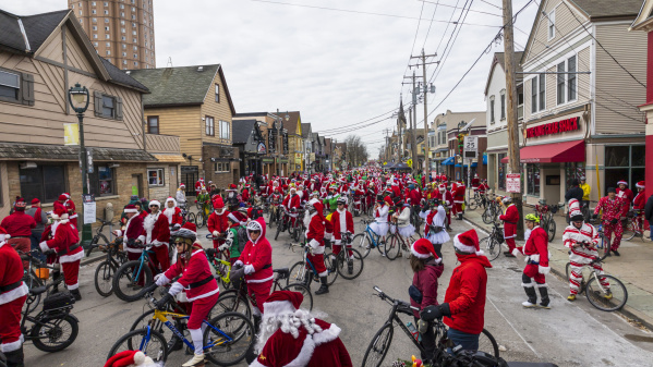 A large group of cyclists dressed in Santa Claus outfits fills a Milwaukee street during a festive holiday bike ride, surrounded by local shops and winter scenery.