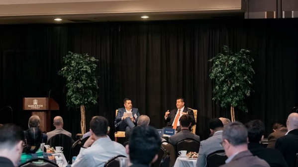 Two speakers sit on stage having a discussion in front of an audience at a formal event. The setting includes a podium, black curtains, and potted trees as stage decor.