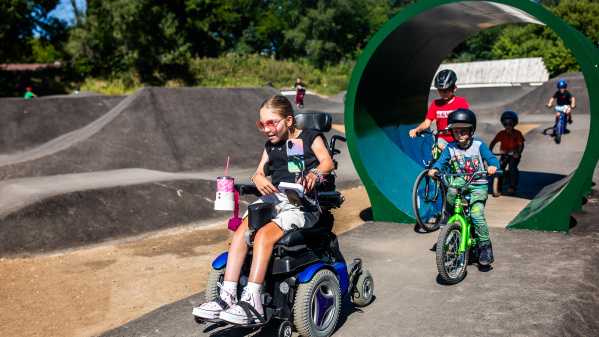 A smiling child using a power wheelchair rides along a pump track at a park, alongside other children on bicycles, with green play structures and trees in the background.