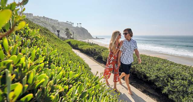 A couple walks along a sunny beach trail in Dana Point, enjoying the warm weather and scenic views.
