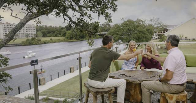 Four adults sit on a riverside terrace, smiling and clinking glasses around a wooden table. Boats pass by on the water below, and trees and buildings are in the background.