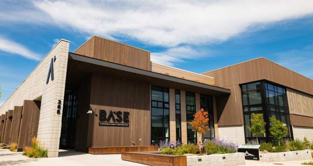 Modern building with large windows and wooden accents, featuring the sign "BASE." Surrounding greenery and blue sky enhance the inviting atmosphere.