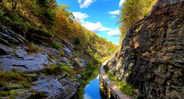 A scenic canal towpath runs alongside still water with dramatic rock walls and bright blue sky overhead.
