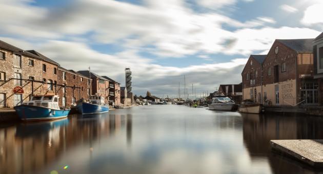 A serene waterfront scene with moored boats on a calm river, flanked by rustic brick buildings under a partly cloudy sky. A peaceful, charming atmosphere.