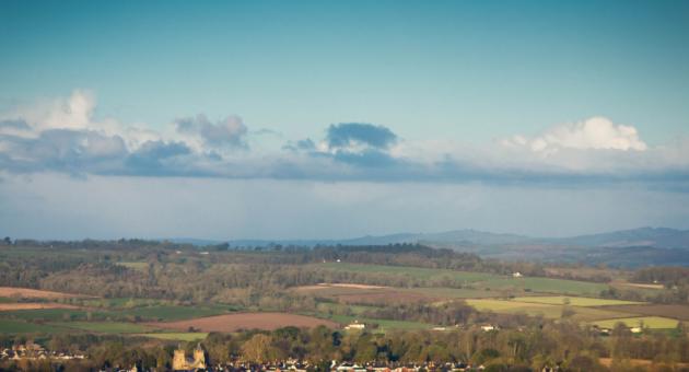 A picturesque rural landscape with patchwork green fields, distant rolling hills, and a small town in the foreground under a vast blue sky with scattered clouds.