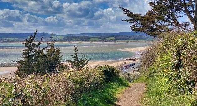 A scenic coastal path leads to a sandy beach, bordered by lush greenery and trees. The sea sparkles under a partly cloudy sky, conveying a serene atmosphere.
