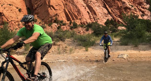Mountain bikers ride through a creek beneath towering red rock cliffs near Cedar City, Utah
