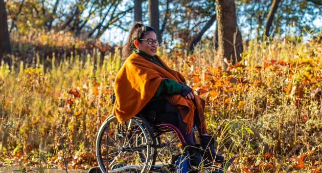 A woman explores a trail while in a wheelchair. The leaves in the background are fall colors.