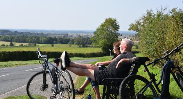 A man and woman sat on a bench in the countryside resting with two bikes next to them,