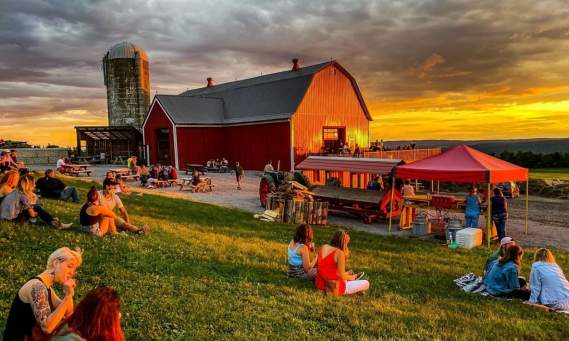 a sunset in back of a red barn with people sitting on the lawn in the foreground