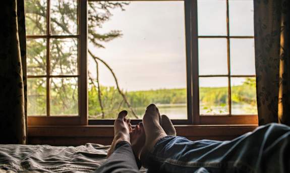 Two people's feet with the window showing view on the lake/wilderness from their cabin