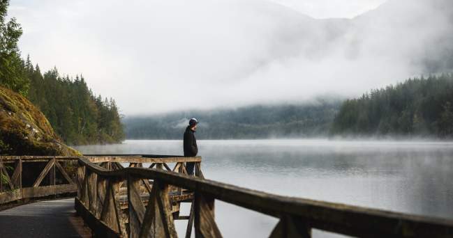 A man stands on a boardwalk looking out over the misty lake.