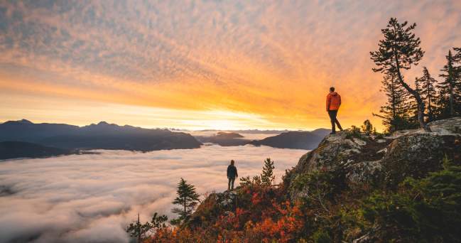 Two people stand on a rocky viewpoint looking at the colours of the sky and the valley below.