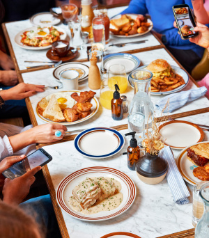 Overhead photo of a group of people gathered around a restaurant table filled with breakfast and brunch dishes. Plates include eggs and toast, bacon, a burger with fries, a sauced entrée, and other shared items, along with drinks like orange juice and water. Several people are holding phones and taking photos of the food.