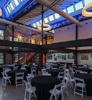 Bloomington event space with round tables covered in black cloths, white chairs, exposed brick walls, large windows, and circular ceiling lights.