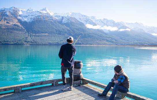 A man fishing off a wharf over a vibrant blue lake and snow covered mountains, while another sits and watches.