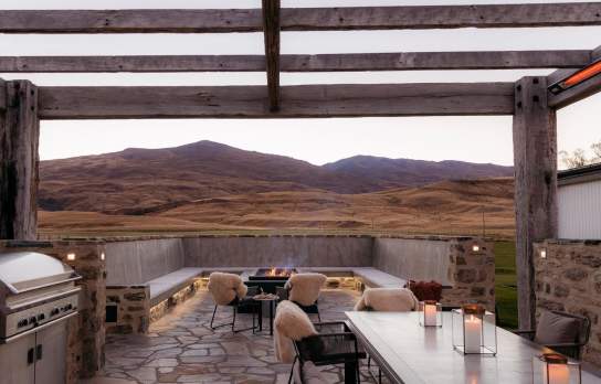 Outdoor dining space at Ben Nevis Station overlooking expansive alpine landscapes, featuring stone flooring, rustic timber beams, and an open-air fireplace setting at dusk.
