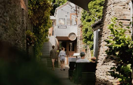 People walking down a side street in Arrowtown with stone buildings and greenery surrounding them
