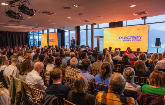A speaker standing on stage in front of a crowd in a conference room with two big screens and views of the mountains in the background