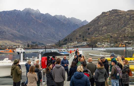 A group of people listening to a person doing a speech on Queenstown Marina with an electric boat and The Remarkable Mountains in the background