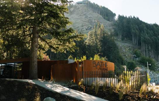 Entrance to a rustic outdoor sauna retreat surrounded by native forest and alpine hillside landscape, featuring timber fencing and natural materials in a secluded setting.