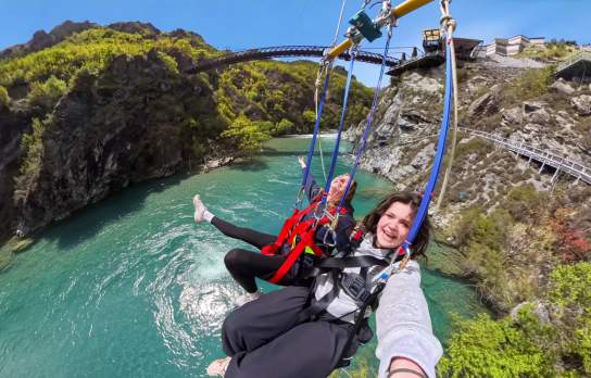 Two people canyon swinging above a turquoise river gorge, suspended by ropes against a rugged cliffside and alpine landscape backdrop.
