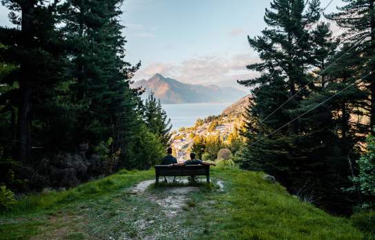 Two people sitting on a chair at the top of a lookout with tall trees in foreground and lake and mountain in background