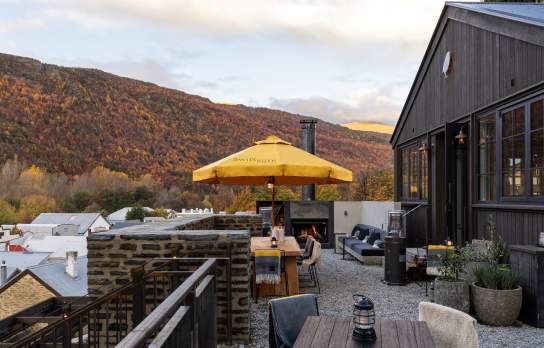 Outdoor terrace at a modern Arrowtown restaurant with mountain views, featuring yellow umbrellas, dining seating, and dark timber exterior in an alpine setting.