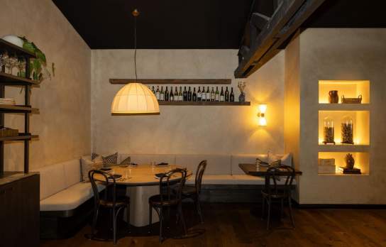 Warm, intimate restaurant interior with softly lit dining tables, curved booth seating, wooden floors, and decorative shelving with ambient lighting and wine display.