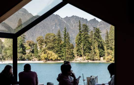 Silhouetted diners seated beside floor-to-ceiling windows overlooking Lake Whakatipu and alpine mountains in a modern restaurant setting.